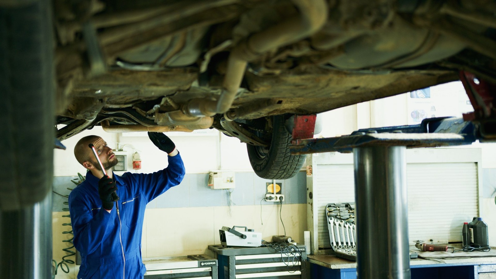 A mechanic using a hand lamp to examine the underside of a raised vehicle
